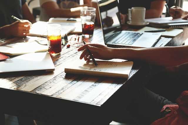 A photo of the hands of people sitting around a table with laptops, notebooks and drinks