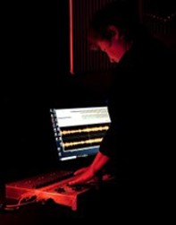 a person stands in the dark illuminated by red light. A computer screen displays soundwaves in the background.