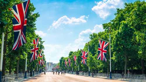 A photograph of The Mall leading towards Buckingham Palace in London. The road is lined with trees  covered by green leaves with Union Jack flags hanging from evenly spaced flagpoles. about halfway down the road is a group of people. Above the sky is blue with some white clouds