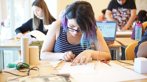 A person in a blue and white striped tank top and glasses draw a straight line on a piece of paper whilst leaning on a light wooden desk. Two more figures are visible behind also crafting.