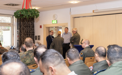 The photo is of a room of people, sitting down and facing away from the camera. Four people are standing up, deep in discussion.