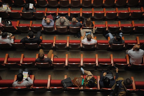 Rows of red seats with wooden armrests seen from above. Several of the seats are occupied by a diverse group of people, some of whom have notepads or tablets on their laps.